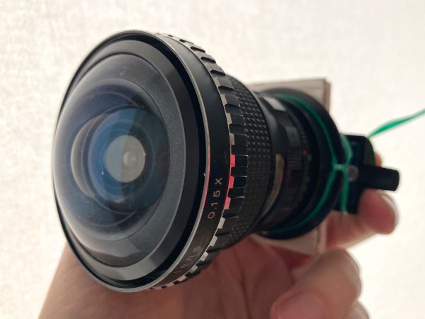 Fisheye-Linse/Adapter vor weißem Hintergrund, gehalten von Hand. Die Linse ist stark gewölbt; am "Hinterteil" klebt ein Stück Pappe und etwas ist mit grünem Gartendraht befestigt.